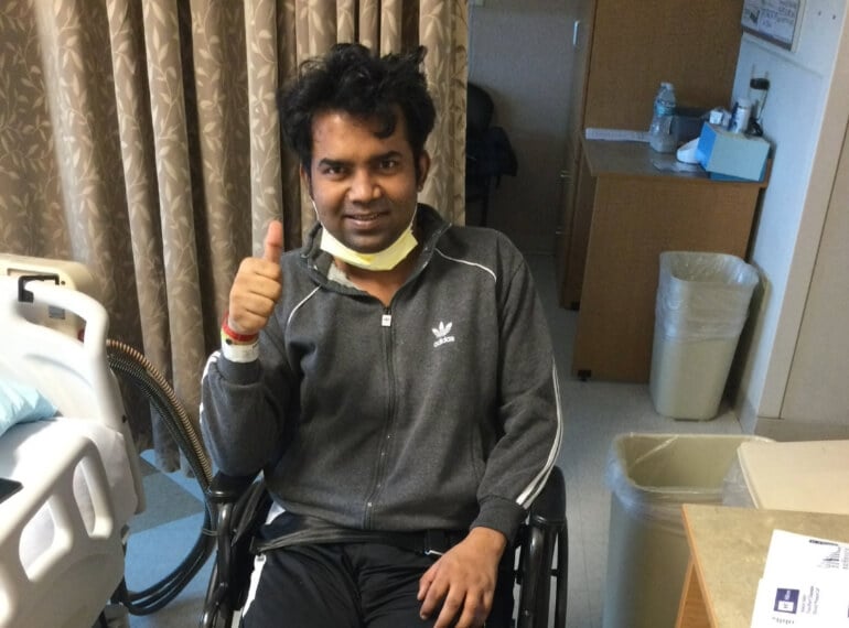 Adesh sitting in a wheelchair in his hospital room giving a thumbs up.