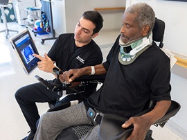 In a rehabilitation clinic, a clinician points to a tablet screen while a seated patient wearing a neck brace and safety belt looks on.