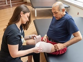 A rehabilitation clinician applies a compression wrap to a patient’s leg while the patient sits on a therapy table.