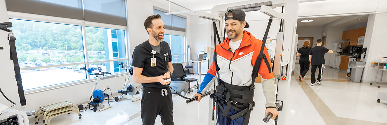 A rehabilitation professional stands beside a patient wearing a body‑weight support harness and holding parallel bars in a therapy gym.