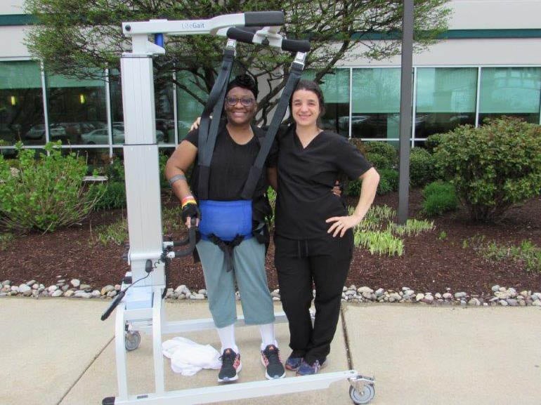 Photo of Linnette Jobson and her therapist walking outside with the help of a harness system at Kessler Institute for Rehabilitation.