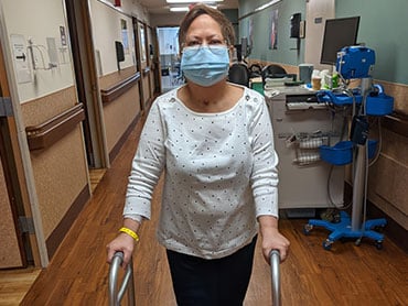 Woman standing outside her hospital room with the aid of a rolling walker wearing a mask.
