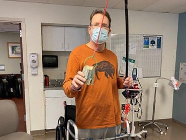 Tom Wolfinger standing in a hospital common area with a mask on holding a fishing pole he used in his occupational therapy.